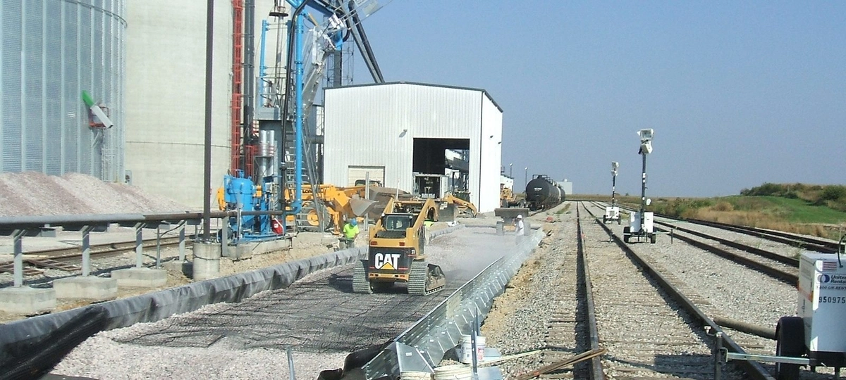 Train tracks run alongside a grain elevator. There is a large white shed between the tracks and ag tanks. A yellow Cat skid loader is driving along the center of the picture.