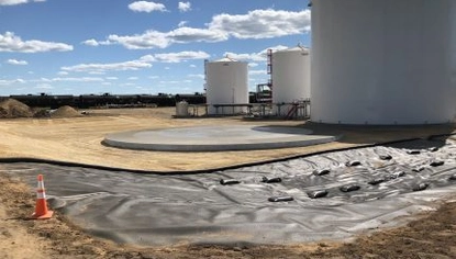 Three tall white container tanks. The one closest is next to a circular base for a tank. Surrounding the area containing the tanks is a black lining on the ground, with a construction cone next to it.