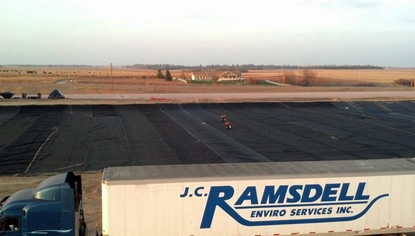 A semi with the JC Ramsdell logo on its trailer is parked in front of a large basin, covered in a black liner. Past the basin is a gravel road, and past that is a ranch-style home.