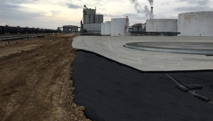 A shallow basin with a circular steel base at its bottom. A black liner covers the front part of the basin. A dirt path runs along the edge of the basin before it dips down. In the background is an ethanol plant with smoke coming out of its smokestack.