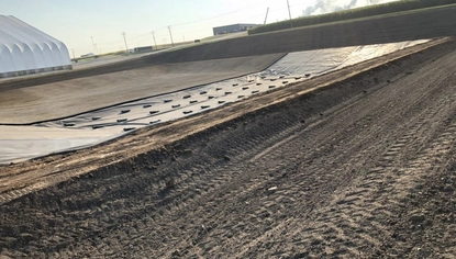 Dirt with tire marks in it. Past the dirt pathway is a basin surrounded by dirt dikes. Part of the basin and the nearby higher ground is covered in a black liner, held down by small rectangular objects.
