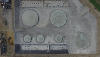 An aerial shot of circular bases sitting in a dirt basin. There are two large bases at the top, with three large bases below, and another large base to their right. On the ground to the left of the basin are stacks of bins and other construction materials.