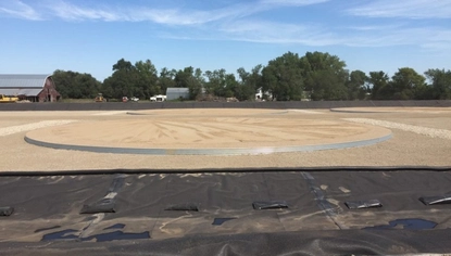 A large, circular steel base sits on a dirt lot. In front of it on the ground is a long black liner.