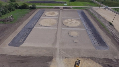 An aerial shot of circular bases, sectioned off in a dirt lot. Two long black liners on the ground extend from the top to the bottom of the area that contains the bases.