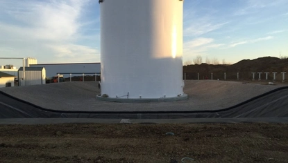 A large white container tank sits in a shallow basin, with parts of the basin and earth above it is covered in a black liner.
