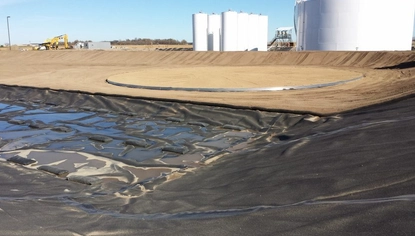 A wide basin with dirt, partially covered by a black liner that runs along the ground and adjoining dikes. Past the liner is a circular steel base. Past the basin on higher ground are large white container tanks.