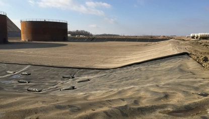 A wide dirt basin. At the far end sit two large, rusted container tanks. Closest to where the picture was taken is a black liner that covers the ground and the adjoining dike.