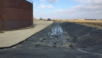 A long black liner runs along the ground, held down by small rectangular black objects. To the left of it is a large, metal container tank that's rusted.