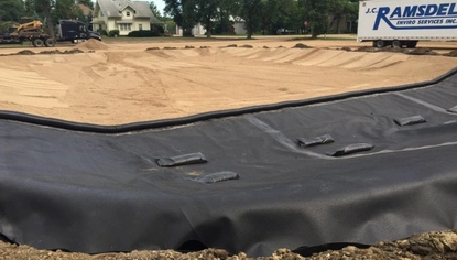 A dirt lot with a slight depression, creating a basin. A trailer with the JC Ramsdell logo on it is to the right of the basin. At the front of the basin is a large black lining that runs along the ground and is held down by black objects.
