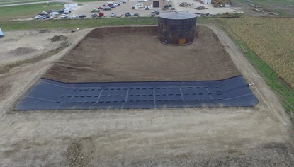 An aerial shot of a dirt basin in a gravel lot. At the far end of the basin is a black container tank. At the closer end is a black covering over the ground and rising slopes around it. In the far background is a large shed with multiple cars parked in the lot surrounding it.