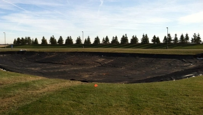 A grassy field with an oval-shaped basin, covered with a black liner. A small orange flag pokes out of the ground before the basin. Past the basin is a line of pine trees on a small hill.