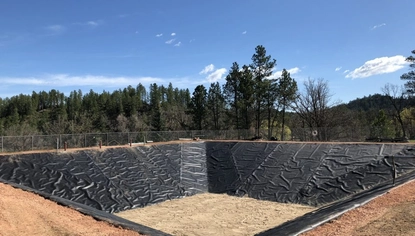 A squared-off basin, covered in a black liner. The base of it is covered in sand. On the level ground beyond it is a chain-link fence, separating the area from a forest.