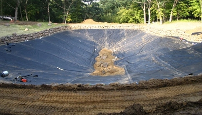 A small, steep basin covered in a black liner. There is a pile of dirt sitting in the middle. A rock wall surrounds most of the basin. There is a lush forest beyond the basin.