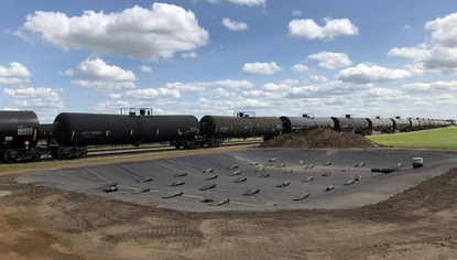 A basin layered with a black liner that's held down by black objects. Next to the dirt basin on the level ground is a train on the train tracks. The sky is blue and scattered with white, fluffy clouds.