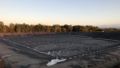 A large basin completely covered with a black liner. The liner is held down by multiple small white objects. On the level ground past the basin is a lush tree line.
