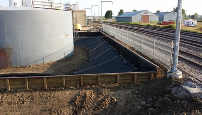 A large gray tank sits in a wooden fenced-off area. There is a chain-link fence separating it from the railroad tracks. On the other side of the tracks sit two long blue sheds.