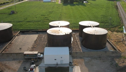 An aerial shot of five large black container tanks. They sit on a dirt lot, but behind them is a grassy field. In front of two of the tanks is a large white shed with a smaller semi parked next to it.