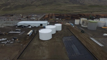 An aerial shot of tanks in a dirt lot. There are more container tanks and industrial buildings and trailers scattered behind the three in the foreground. In the background is a mountain range.