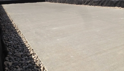 Close-up of a concrete slab. The sun is lighting up the surface while the rocks that line it are in the shade. Tall maroon container tanks are in the background with a pickup truck parked in front of them.
