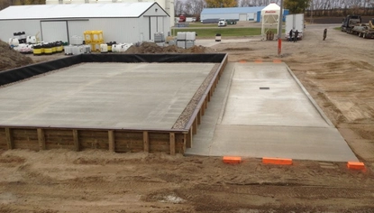 Two concrete slabs sit on a dirt lot. One is sectioned off by a short wooden wall with rocks lining the inside of the fence. The other slab is on its own, with bright orange blocks sitting at opposite ends of it. In the background is a large white shed and a few people walking around on the dirt lot.