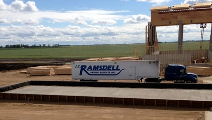 A concrete rectangular slab on a dirt lot, fenced off by a short, black enclosure. A semi with the JC Ramsdell logo on its trailer is parked right beyond it. In the background and to the right is a large structure under construction. The scene takes place in front of a grassy field.