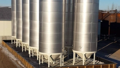 Multiple tall and skinny silver container tanks held up by short white support beams. The tanks sit in a fenced-off area.