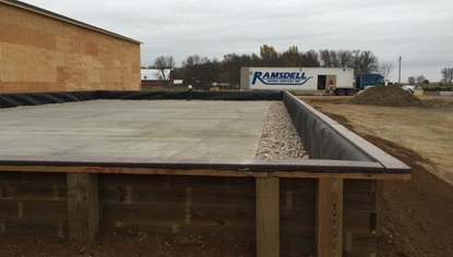 A short wooden wall contains a concrete slab with rocks lining the rims. Next to the patch is a structure with walls made of oriented strand board. A semi with a JC Ramsdell logo on the trailer is parked in the background.