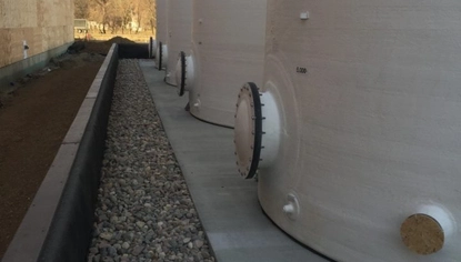 A close-up of the back ends of white container tanks. There is a port at the bottom of each one that's closed off with the lids screwed in. The tanks sit on concrete with a patch of rocks going around the perimeter of the slab. There is a building made of oriented slab boards on the opposite end of the tanks.