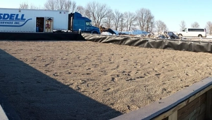 A fenced-off area filled with dirt. In the background of the enclosed area is a blue semi with a trailer that has the JC Ramsdell logo on it. It's winter, so there are small patches of snow with larger piles of snow in the far background. A white 18-passenger van is parked next to one of the snow piles.