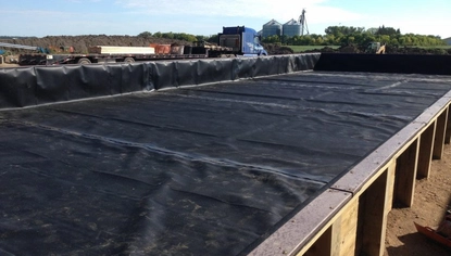 A fenced-off area, layered in a large black liner covering. A blue semi is parked in the background.