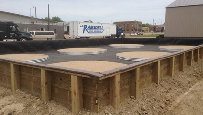 Circular wooden slabs are placed in a fenced-off area. The enclosure is lined with a large black tarp. Behind it sits a semi with the JC Ramsdell logo on its trailer, along with other semis and cars.