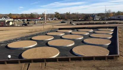 Multiple flat wooden rings sit on a black tarp, which is enclosed by a short wooden wall in a dirt lot. A gas station is in the background. The sky is blue with a few white clouds.