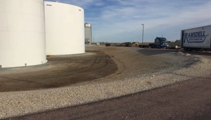 Two large container tanks sit in a shallow dirt basin. Along the perimeter of the basin is gravel with two semi trucks parked next to it.