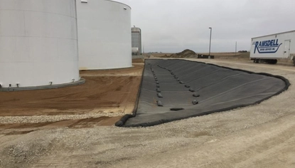 Two white tanks sit in a shallow basin. Next to the tanks is a long, black liner that covers the ground and the dike on the right side of the basin. A semi-truck trailer with the JC Ramsdell logo on it is parked to the right of the basin.
