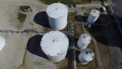 An aerial photo of five white container tanks placed in a gravel lot. There are pipelines running between them and connecting to other structures on the right and left sides of the photo.