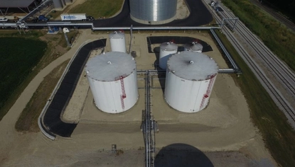 An aerial picture of white container tanks sitting in a sandy basin. Black liners cover parts of the dike built around the basin. There is a larger steel grain bin in the background, also surrounded by a black liner. There are industrial buildings and equipment to the left of the grain bin. Railroad tracks run alongside the lot on its right side.
